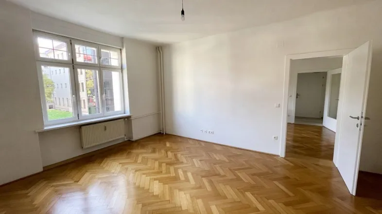 Helles Zimmer mit Fischgrätparkett und großen Fenstern, das viel Tageslicht hereinlässt.