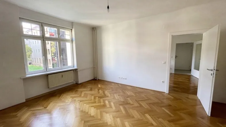 Helles Zimmer mit Fischgrätparkett und großen Fenstern, bereit zur individuellen Gestaltung und Nutzung.