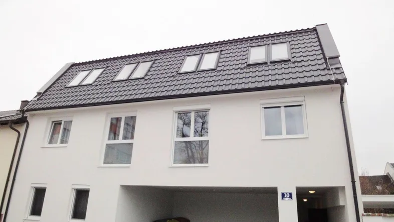 Weißes Mehrfamilienhaus mit dunklem Mansarddach und mehreren Dachfenstern, inklusive Carport.