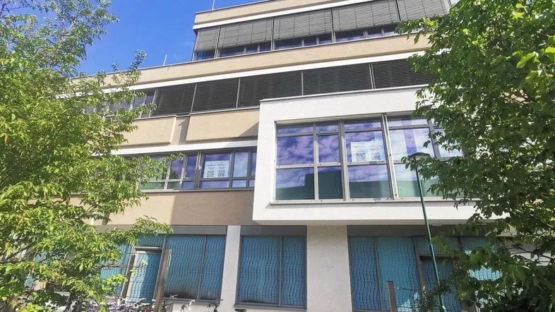 Modernes Bürogebäude mit großen Fenstern und Jalousien, umgeben von grünen Bäumen und Fahrradstellplätzen.