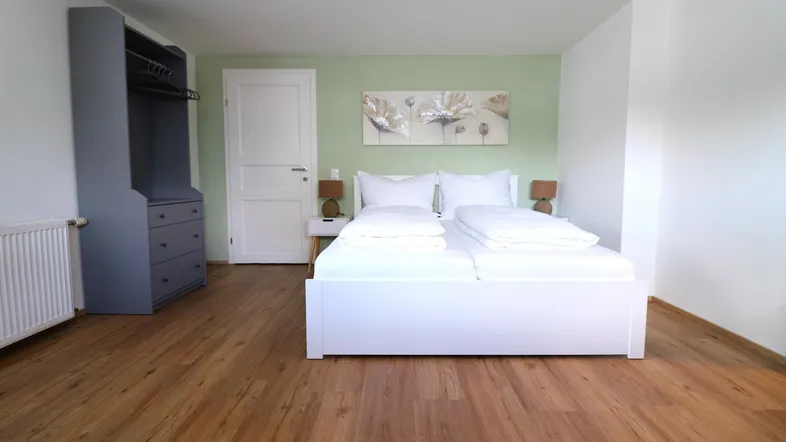 Helles Schlafzimmer mit Doppelbett, Holzfußboden und einer Wand in Pastellgrün.