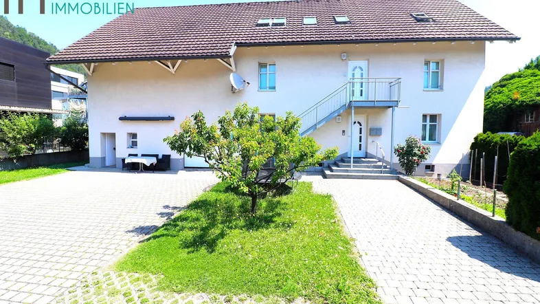 Vorderansicht des Hauses mit gepflasterter Einfahrt, kleinem Baum und Bergblick im Hintergrund.