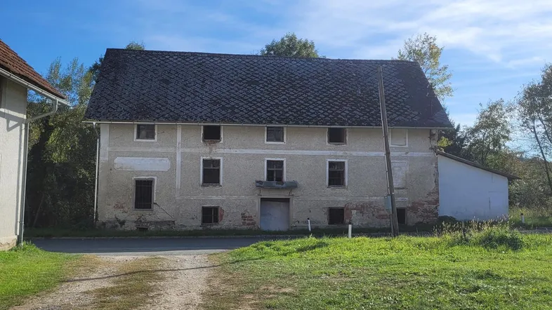 Altes, verputztes Gebäude mit dunklem Dach und einer unbefestigten Zufahrt auf dem Land.