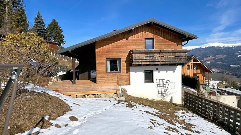 Modernes Holzhaus mit weißer Fassade und Bergblick, umgeben von Bäumen und Schnee.