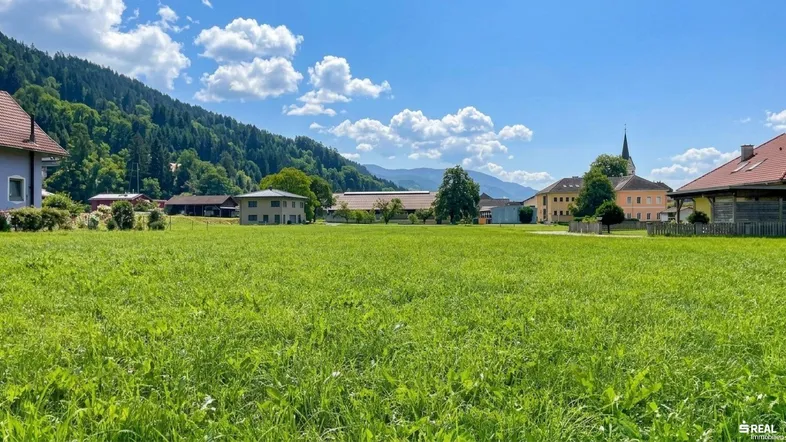 Weitläufige grüne Wiese mit Blick auf umliegende Häuser, Berge und einen Kirchturm unter blauem Himmel.