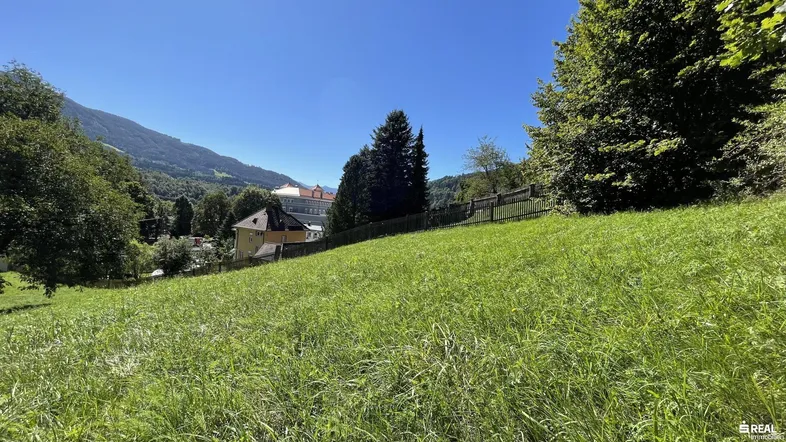 Grüne Wiese mit Blick auf die umliegende Berglandschaft und entfernte Gebäude unter blauem Himmel.