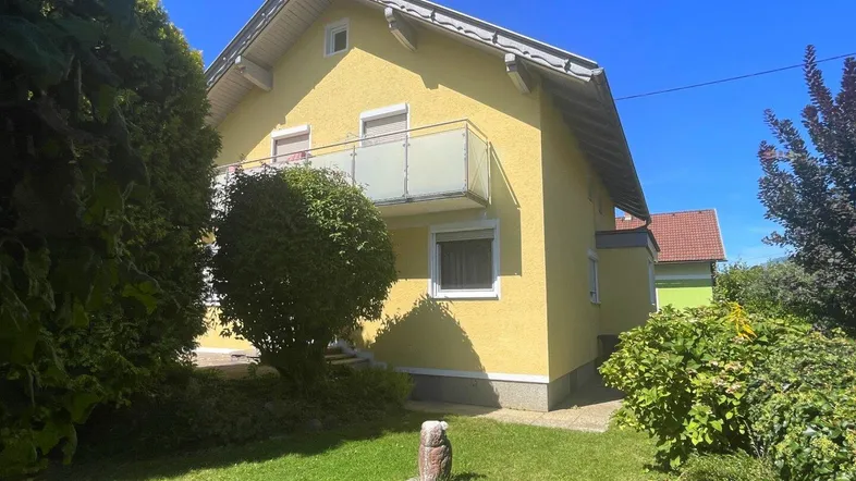 Gelbes Einfamilienhaus mit Balkon und gepflegtem Vorgarten unter blauem Himmel.