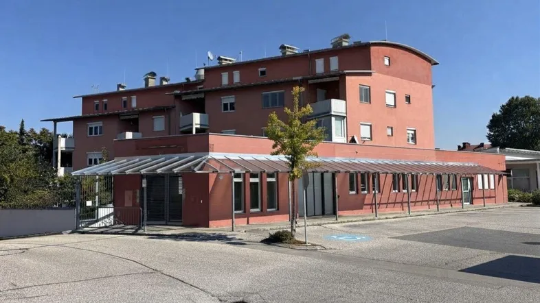 Mehrstöckiges Gebäude mit roter Fassade, überdachtem Eingangsbereich und Parkplätzen davor unter blauem Himmel.
