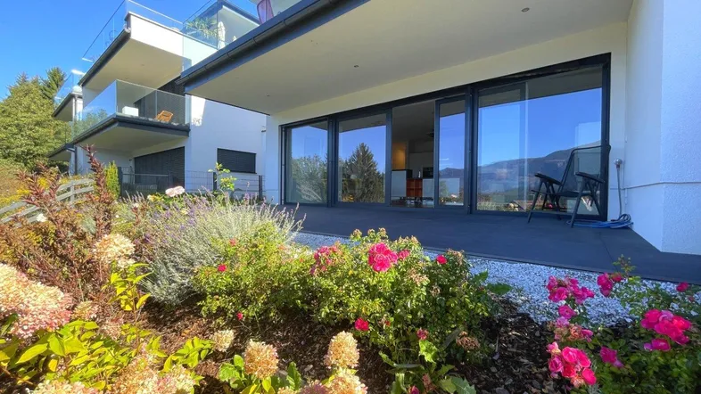 Moderne Wohnanlage mit großer Terrasse, blühendem Garten und weitem Blick auf die Berge und den See.