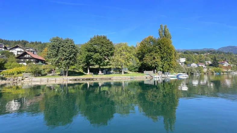 Malerischer Seeblick mit grünen Bäumen und Häusern am Ufer unter blauem Himmel.