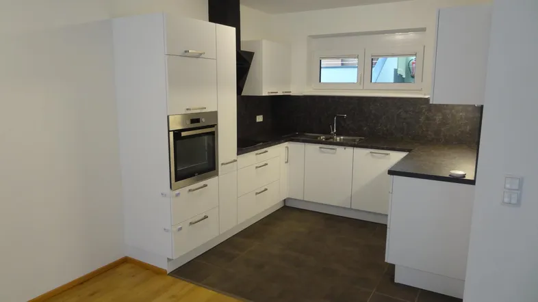 Moderne Einbauküche mit weißen Schränken, dunkler Arbeitsplatte, integriertem Backofen und Fenster.