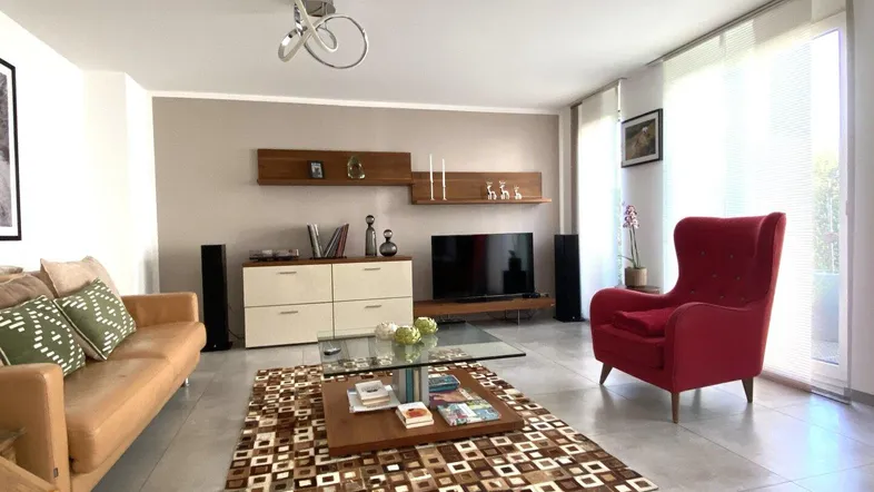Helles Wohnzimmer mit beigem Sofa, rotem Sessel und großen Fenstern.