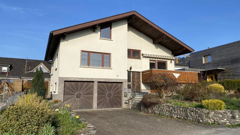 Einfamilienhaus mit Satteldach, Doppelgarage, Balkon und gepflegtem Vorgarten unter blauem Himmel.