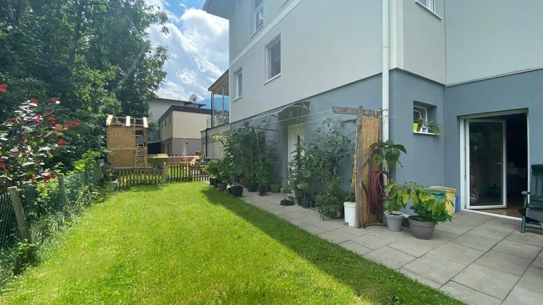 Grüner Gartenbereich mit Rasenfläche, kleiner Terrasse und bepflanzten Beeten neben dem Haus.