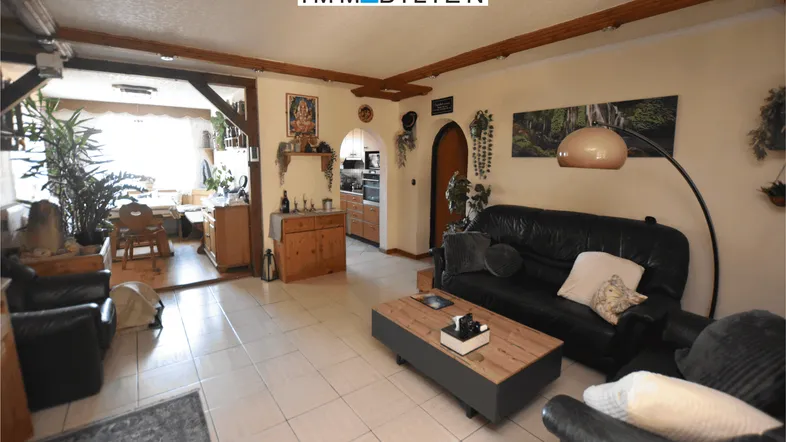 Gemütliches Wohnzimmer mit dunklen Ledersofas, Fliesenboden und offenem Übergang zum Essbereich.