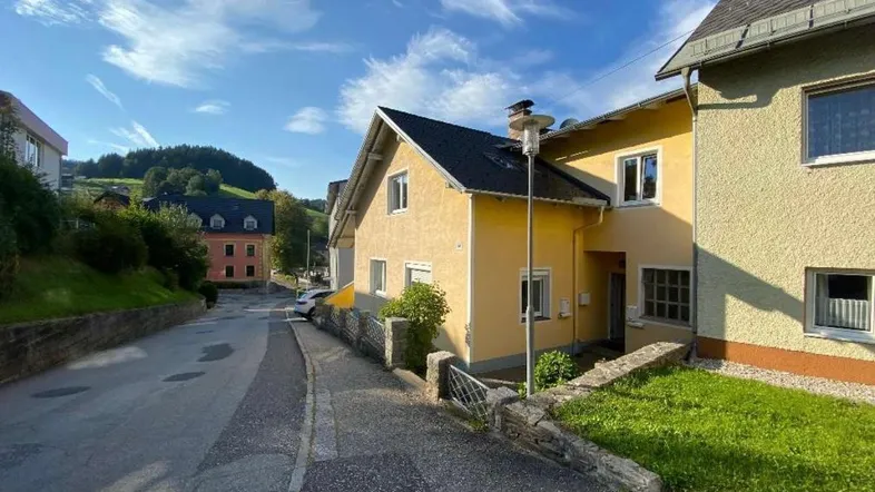 Gelbes Wohnhaus an einer leicht ansteigenden Straße in einer ländlichen Umgebung mit grünen Hügeln.