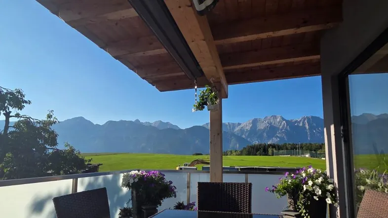 Sonniger Balkon mit Holzdach, Sitzgelegenheiten und beeindruckendem Bergblick über grüne Felder.