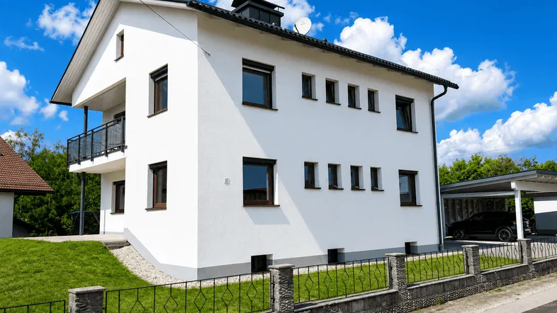 Weißes Einfamilienhaus mit Balkon, Vorgarten und Carport unter blauem Himmel.