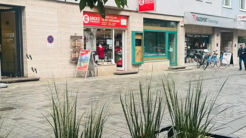 Blick auf eine belebte Geschäftsstraße mit verschiedenen Ladenfronten und Pflanzen im Vordergrund.