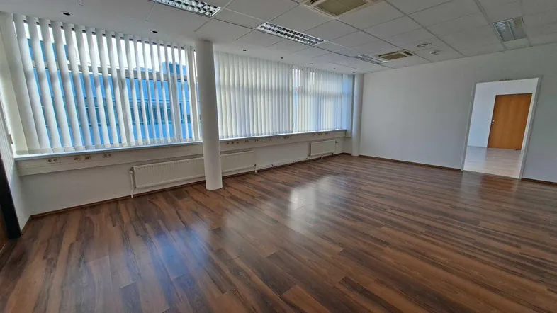 Geräumiges, leeres Büro mit großen Fenstern, Jalousien und dunklem Laminatboden unter einer Rasterdecke.