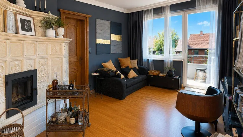 Gemütliches Wohnzimmer mit traditionellem Kachelofen, Holzboden und Zugang zum Balkon.