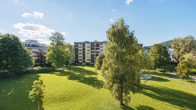 Weitläufige Grünfläche mit Bäumen und Mehrfamilienhäusern im Hintergrund unter blauem Himmel.