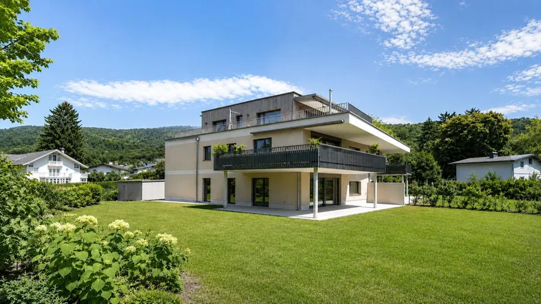 Modernes Mehrfamilienhaus mit großem Garten, Balkonen und Bergblick unter blauem Himmel.