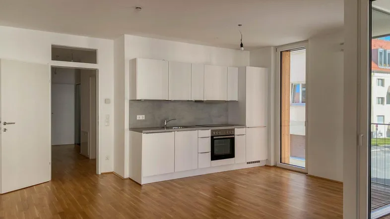 Heller offener Wohnbereich mit integrierter Küchenzeile, Parkettboden und Zugang zum Balkon.