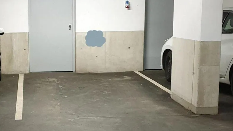 Tiefgaragenstellplatz mit Betonboden, weißen Wänden, grauen Türen und sichtbaren Installationen an der Decke.