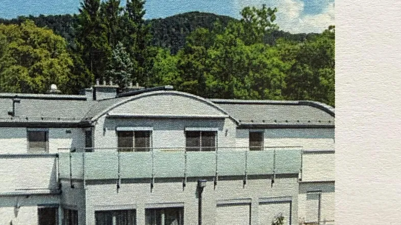 Mehrfamilienhaus mit Balkonen und Blick auf bewaldete Hügel unter blauem Himmel.