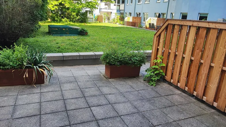 Gepflasterte Terrasse mit Pflanzenkübeln und Zugang zu einem grünen Gemeinschaftsgarten.
