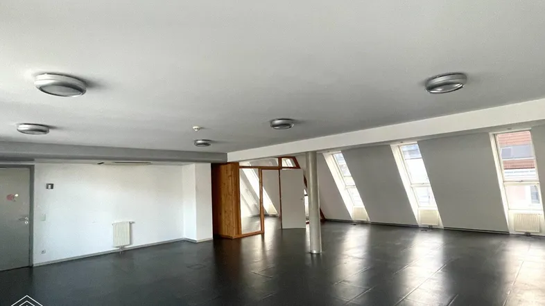 Großer, heller Büroraum mit dunklem Fliesenboden, schrägen Dachfenstern und einer sichtbaren Betonsäule.