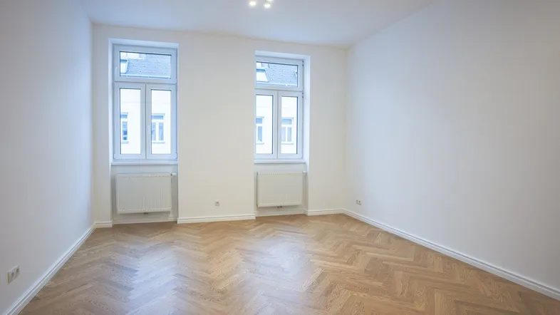 Helles, leeres Zimmer mit Fischgrätparkettboden und zwei Fenstern, bereit zur individuellen Gestaltung.
