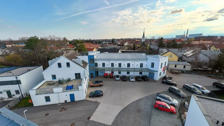 Luftaufnahme des Gebäudekomplexes mit Parkplätzen und umliegender Bebauung unter blauem Himmel.