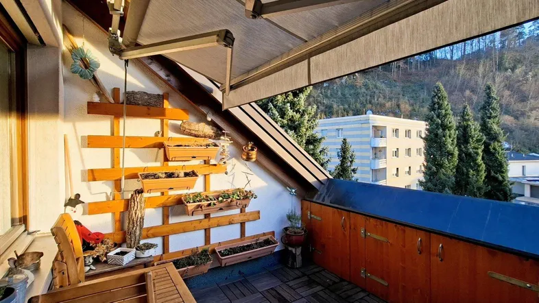 Geräumiger Balkon mit Holzboden, Markise und Blick ins Grüne sowie auf die umliegenden Gebäude.