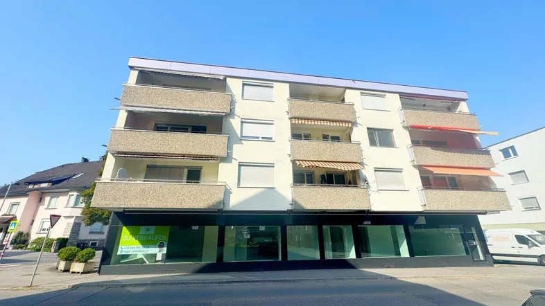 Mehrstöckiges Wohngebäude mit beige gestrichener Fassade, mehreren Balkonen und Rollläden an den Fenstern.