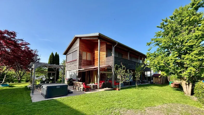 Geräumiger Garten mit Holzhaus, Terrasse, Whirlpool und Pergola unter blauem Himmel.