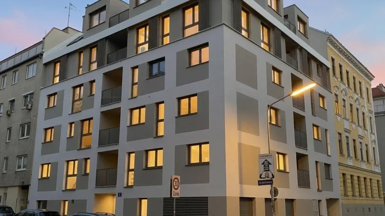 Modernes Mehrfamilienhaus mit beleuchteten Fenstern in der Dämmerung, umgeben von städtischer Architektur.