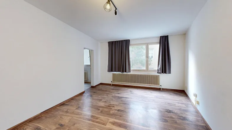 Helles Zimmer mit Holzboden, weißen Wänden und einem Fenster mit dunklen Vorhängen.