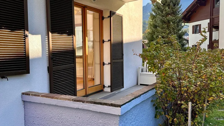 Kleiner Balkon mit Holzfensterläden und Markise, umgeben von grüner Vegetation.