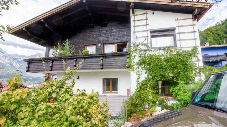 Traditionelles Haus mit Holzbalkon und üppiger Begrünung, umgeben von einer bergigen Landschaft.