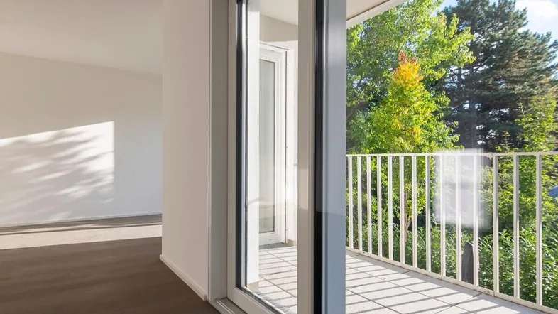Helles Zimmer mit dunklem Holzboden und Zugang zu einem Balkon mit Grünblick.