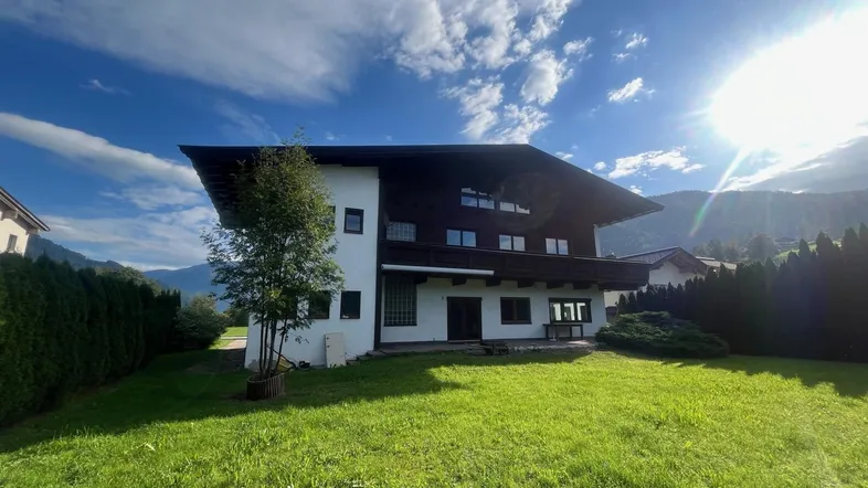 Hausfassade im alpinen Stil mit großem Rasen und Bergblick unter blauem Himmel.