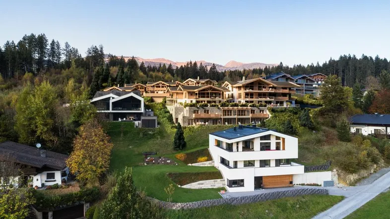Panoramablick auf eine Ansammlung von modernen und traditionellen Häusern in einer bewaldeten Hanglage mit Bergblick.
