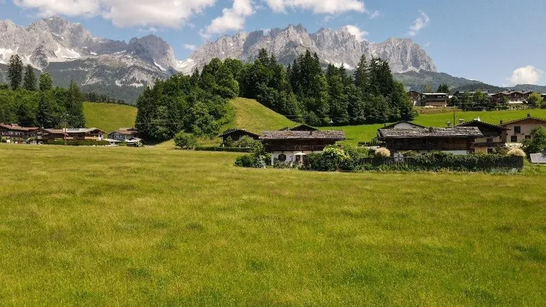 Grüne Wiesen mit traditionellen Häusern und majestätischem Bergpanorama unter blauem Himmel.