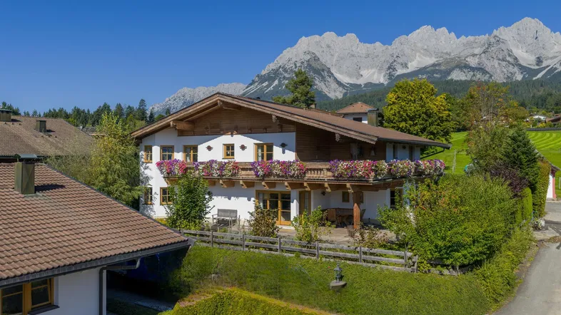 Traditionelles Haus im Alpenstil mit blühenden Balkonkästen und beeindruckendem Bergblick unter blauem Himmel.