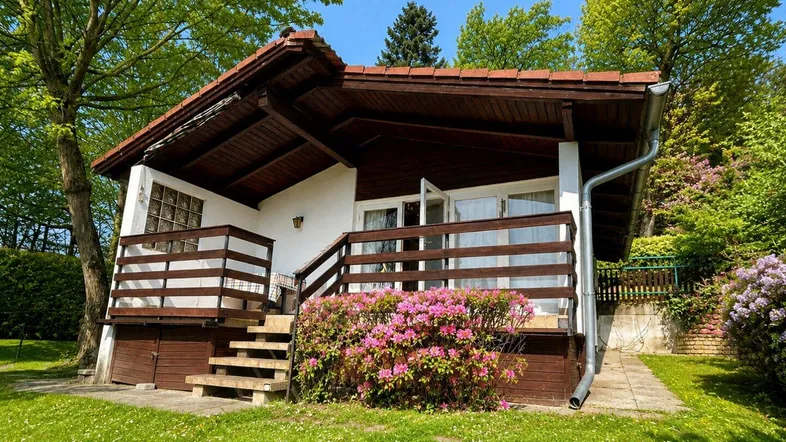 Charmantes kleines Haus mit Holzveranda und üppiger grüner Umgebung unter blauem Himmel.