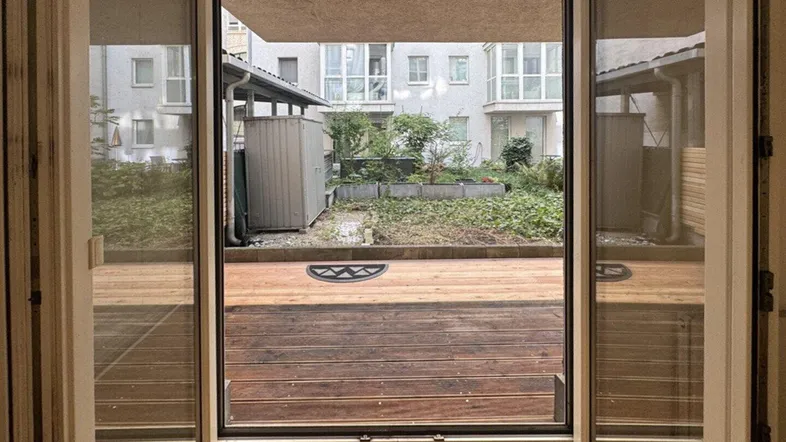 Blick aus einem hellen Raum auf eine Holzterrasse und einen begrünten Innenhof.
