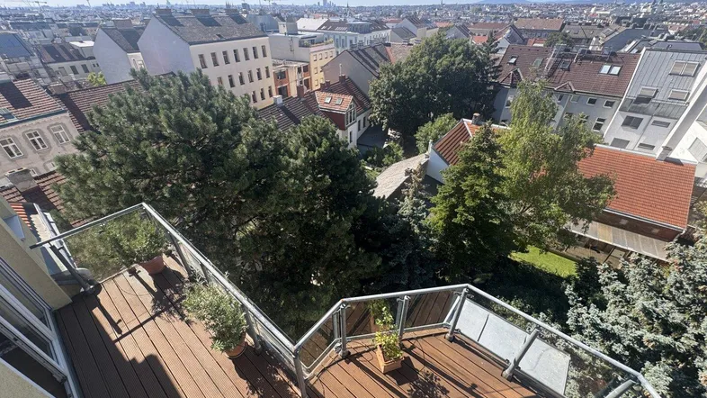 Geräumiger Balkon mit Holzboden, Glasgeländer und weitem Blick über die Stadt und grüne Bäume.