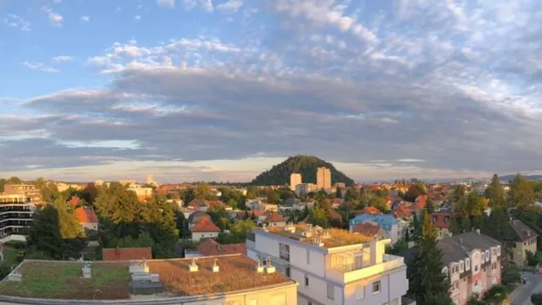 Panoramablick über die Stadt mit grünen Dächern und einem bewaldeten Hügel im Hintergrund bei Sonnenaufgang.